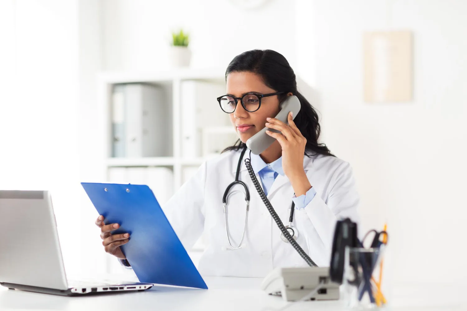 doctor in a white coat and glasses talking on a desk phone while reviewing a clipboard, with a laptop in front of her